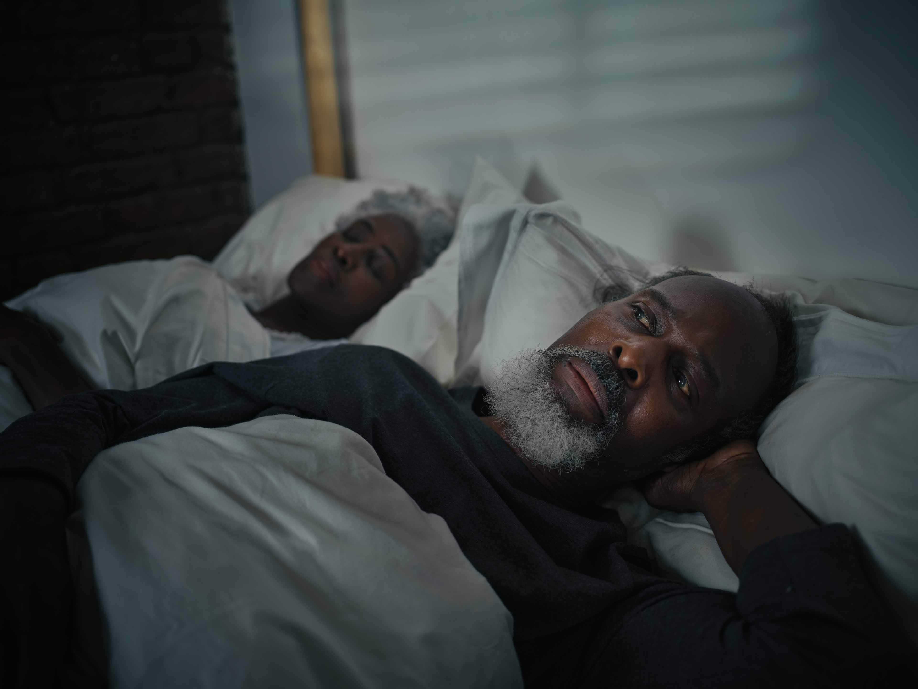 Mature man laying awake in bed while his wife is sound asleep next to him