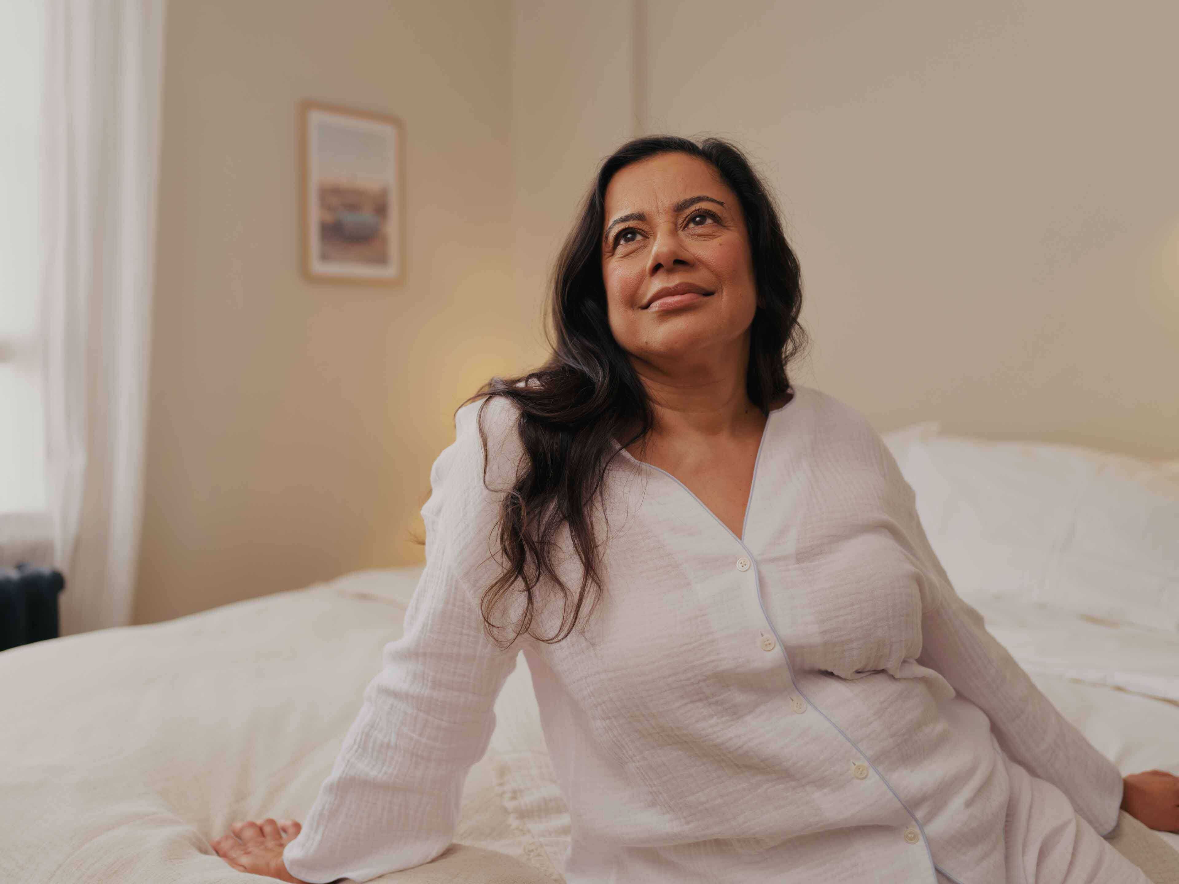 Mature woman with long dark hair sitting at the edge of her bed wearing pyjamas looking outward smiling and looking well rested
